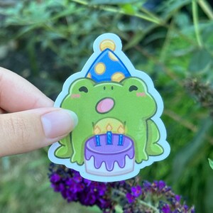 May include: A green frog sticker wearing a blue party hat with yellow polka dots. The frog is holding a purple birthday cake with three lit candles. The sticker is held in front of a blurred green background.