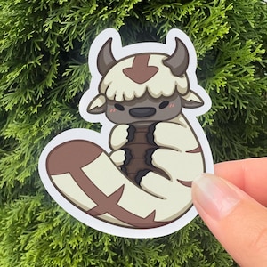 May include: A white and brown cartoon sticker of Appa, the flying bison from Avatar: The Last Airbender, curled up in a ball. Appa has a brown nose, a brown arrow on his forehead, and a white fluffy mane.