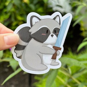 May include: A cute cartoon raccoon holding a blue sword with a brown handle. The raccoon has a gray body, black eyes, and a black and white tail.