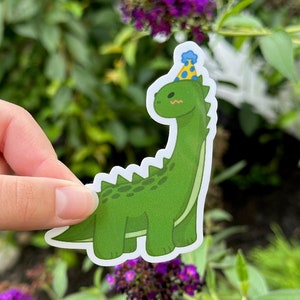 May include: A green dinosaur wearing a blue and yellow party hat. The dinosaur has a long neck and a friendly face.