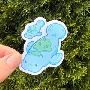 May include: A die-cut sticker featuring a large light blue manatee with a patch of green seaweed on its back and a smaller matching baby manatee floating above it. Both creatures have simple facial features, pink cheeks, and a white border.