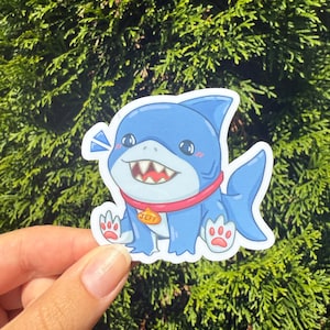 May include: A blue shark sticker with a red collar and the name "JEFF" on a tag. The sticker has a cartoon design with a smiling shark and is held against a green leafy background.