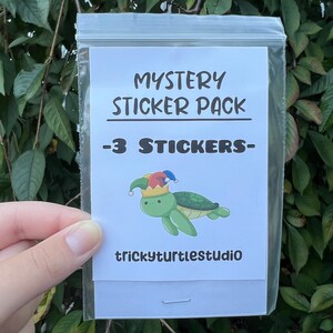 May include: A white plastic bag with a white card inside. The card has black text that says "Mystery Sticker Pack - 3 Stickers -" and a cartoon image of a green turtle wearing a colorful jester hat. The text "trickyturtle studio" is at the bottom of the card.