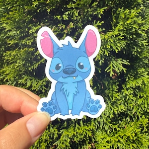 May include: A blue sticker featuring a cartoon character with large ears, big eyes, and a small mouth. The character is sitting down with its paws visible. The sticker has a white border and is set against a backdrop of green foliage.