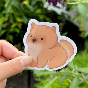 May include: A sticker featuring a cartoon-style Chow Chow dog. The dog is depicted in shades of brown and tan with a white outline. The sticker has a cute, friendly appearance, with rosy cheeks and a small nose.