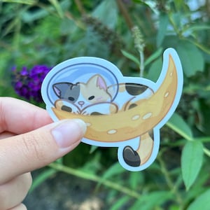 May include: A white, black, and orange cat wearing a space helmet is sleeping in a crescent moon. The cat is curled up in the moon, with its tail wrapped around its body. The moon is a light yellow color with a white outline.