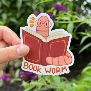 May include: A sticker of a pink worm wearing glasses and reading a red book with gold accents. The sticker says "BOOK WORM" in orange letters.
