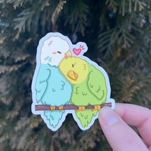 May include: A sticker featuring two cartoon parakeets, one blue and one green, with a small pink heart above them. The parakeets are perched on a branch and appear to be cuddling.