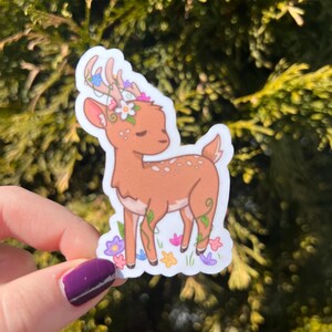 May include: A brown deer with white spots and antlers adorned with flowers. The deer is standing in a field of flowers, including pink, purple, and yellow.