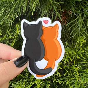 May include: A sticker featuring two cats, one black and one orange, with a small red heart above them. The cats are facing each other and appear to be cuddling.