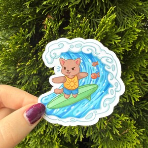 May include: A die-cut sticker featuring a brown cartoon cat with a determined expression, wearing a yellow sun-print tank top and blue shorts, surfing on a green surfboard inside a large blue ocean wave with two small orange fish nearby.