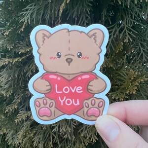 May include: A brown teddy bear holding a red heart that says "Love You" in white text. The bear has pink paw pads and a light blue outline.