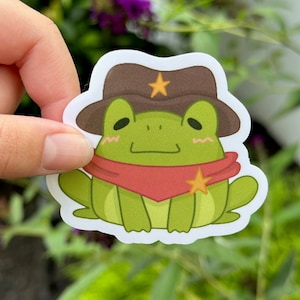 May include: A green frog wearing a brown cowboy hat with a yellow star and a red bandana with a yellow star. The frog is sitting with its legs crossed.
