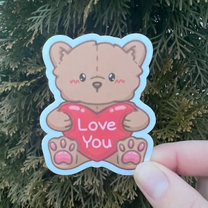 May include: A brown teddy bear holding a red heart that says "Love You" in white text. The bear has pink paw pads and a light blue outline.