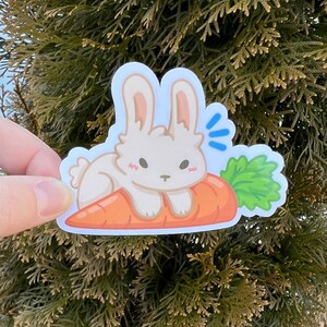 May include: A white bunny rabbit sticker with pink cheeks is sitting on a bright orange carrot with green leafy tops. The bunny has a surprised expression with its mouth open and its eyes wide.