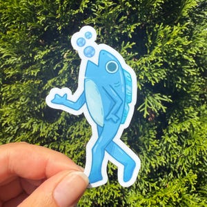 May include: A blue sticker featuring a cartoon fish with legs, walking and blowing bubbles. The fish has a light blue belly and a dark blue outline. The sticker is held against a background of green foliage.