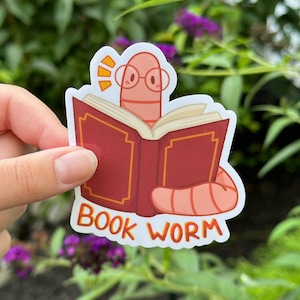 May include: A sticker of a pink worm wearing glasses and reading a red book with gold accents. The sticker says "BOOK WORM" in orange letters.