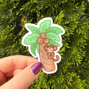 May include: A die-cut sticker featuring a cartoon illustration of a brown monkey clinging to a tan palm tree trunk. The tree has green palm fronds at the top and three brown coconuts clustered in the center. The design has a white border outline.