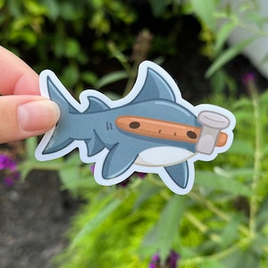 May include: A blue shark sticker with a brown nose and a white cup. The shark is holding the cup with its mouth.