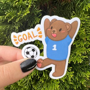 May include: A brown bear wearing a blue jersey with the number "1" on it, holding a soccer ball. The bear is standing on a white background with the word "GOAL" in orange letters.