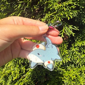 May include: A light blue shark keychain with a nurse's hat and bandages. The keychain has a silver clasp and a colorful iridescent clip. The shark has a cute, cartoonish design and is held against a background of green foliage.