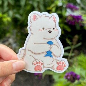 May include: A white polar bear sticker with pink cheeks and paws, holding a blue fish. The sticker is on a white background.