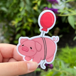 May include: A pink pig sticker with a red balloon tied to its tail. The pig has a cute cartoon face and is holding onto the balloon string.