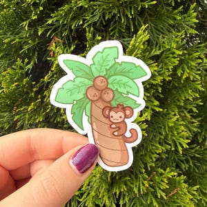 May include: A die-cut sticker featuring a cartoon illustration of a brown monkey clinging to a tan palm tree trunk. The tree has green palm fronds at the top and three brown coconuts clustered in the center. The design has a white border outline.