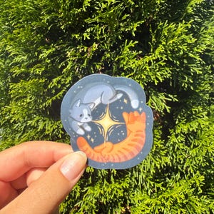 May include: A sticker featuring two cats, one gray and one orange, in a celestial design. The gray cat is lying on its back, while the orange cat is curled up. The background is dark blue with stars and a bright yellow starburst.