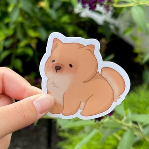 May include: A sticker featuring a cartoon-style Chow Chow dog. The dog is depicted in shades of brown and tan with a white outline. The sticker has a cute, friendly appearance, with rosy cheeks and a small nose.