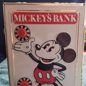 Peut inclure: Une tirelire en métal vintage de Mickey Mouse avec un schéma de couleurs rouge et blanc. La tirelire a un cadenas à combinaison avec un bouton rouge et les mots "Mickey's Bank" imprimés en blanc sur un fond rouge.