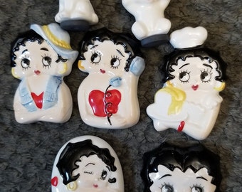 Puede incluir: Conjunto de siete figuras de cerámica que representan a un personaje de dibujos animados con cabello negro y ojos grandes. Las figuras están en varias poses y atuendos, incluyendo un vestido rojo, una camisa azul y un gorro de chef blanco. También hay dos figuras de perros blancos.