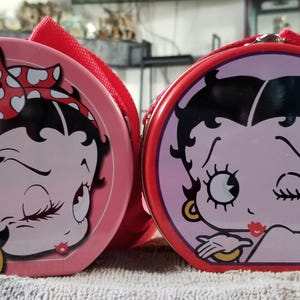 以下が含まれることがあります： 2つの丸いピンク色のベティ・ブープをテーマにした化粧品ケース。片方は白いハートの赤いリボンを着けたベティ、もう片方はウィンクしてキスを送っています。どちらも赤いハンドルが付いています。