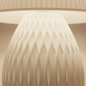 May include: A white, textured lamp base with a geometric diamond pattern. The lamp base is cone-shaped and sits beneath a white lampshade. The lamp is lit, casting a soft glow.