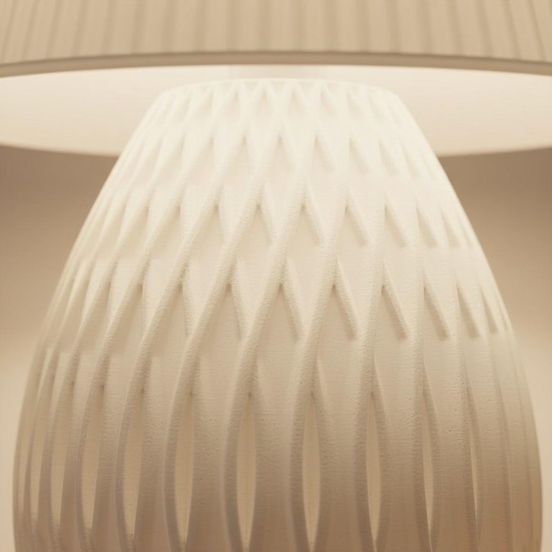 May include: A white, textured lamp base with a geometric diamond pattern. The lamp base is cone-shaped and sits beneath a white lampshade. The lamp is lit, casting a soft glow.