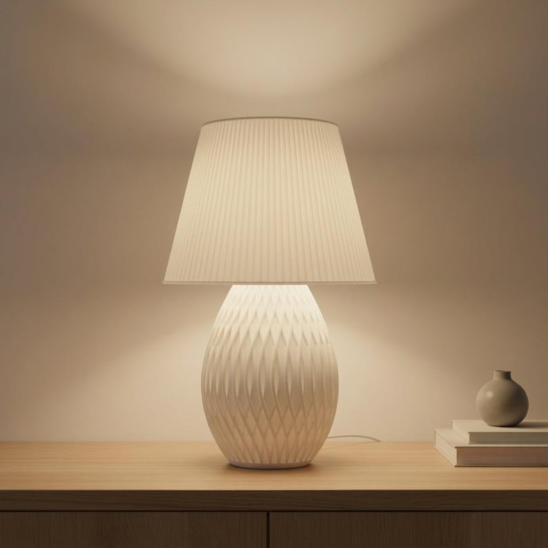May include: A white table lamp with a pleated lampshade and a textured, egg-shaped base. The lamp is on a wooden surface. A small vase and books are also on the surface.
