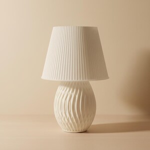 May include: A white ceramic table lamp with a pleated lampshade. The base has a textured, swirling design. The lamp is a neutral color and would fit in many home decor styles.