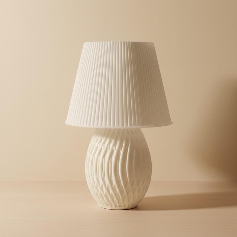 May include: A white ceramic table lamp with a pleated lampshade. The base has a textured, swirling design. The lamp is a neutral color and would fit in many home decor styles.