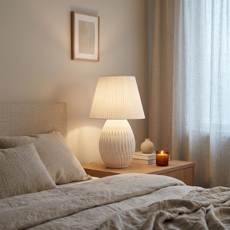 May include: A white table lamp with a pleated lampshade sits on a wooden nightstand. The lamp has a textured base. A framed picture hangs on the wall above the bed. The bed has beige bedding and pillows. A lit candle and a small vase are on the nightstand.