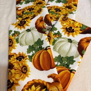 Könnte beinhalten: Vier gefaltete weiße Stoffservietten mit einem sich wiederholenden Muster aus Sonnenblumen, Kürbissen und grünen Blättern. Die Kürbisse sind orange und weiß, und die Sonnenblumen sind gelb und braun. Die Servietten sind wahrscheinlich für eine herbstliche oder erntezeitliche Tischdekoration.