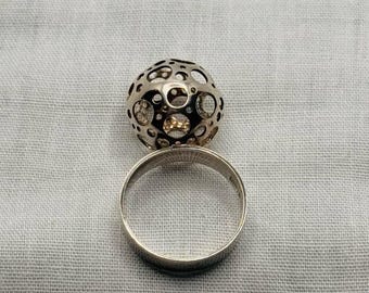 May include: A silver ring with a hollow sphere design. The sphere is covered in circular cutouts, revealing a gold-colored interior. The ring band is a simple, polished silver. The jewelry is set against a neutral, textured background.