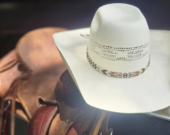 May include: A white cowboy hat with a brown leather band and a beaded trim. The hat is sitting on a brown leather saddle.