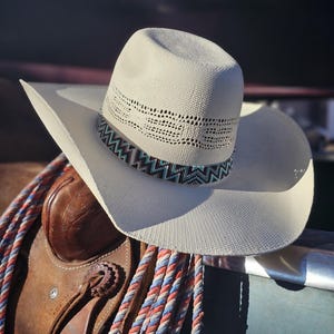 Peut inclure: Un chapeau de cowboy en paille blanche avec un bandeau de perles turquoise et marron. Le chapeau est posé sur une selle en cuir marron avec une corde rouge, blanche et bleue.