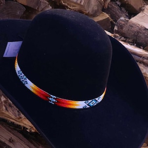 Może przedstawiać: Czarny kowbojski kapelusz z ozdobnym paskiem z koralików w geometrycznym wzorze w odcieniach bieli, pomarańczy, czerwieni i błękitu.