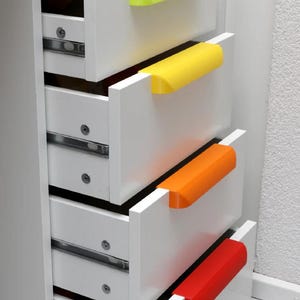 Puede incluir: Un mueble blanco con cinco cajones, cada uno con un tirador de diferente color: verde, amarillo, naranja y rojo. Los cajones están parcialmente abiertos, revelando el interior y las guías metálicas. El mueble está contra una pared blanca.