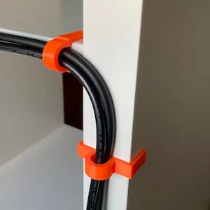 Puede incluir: Clips de cable naranja que aseguran cables blancos y negros a una superficie blanca. Los clips son de plástico y están diseñados para mantener los cables organizados. Los cables están doblados.