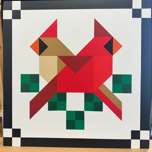 May include: A barn quilt with a geometric design of two red cardinals with brown beaks and black eyes, perched on a green leafy branch.