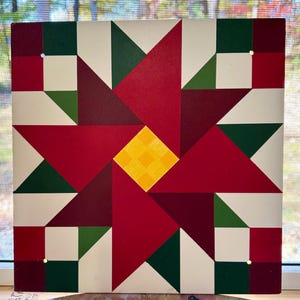 May include: A red, green, white, and yellow barn quilt with a starburst design. The quilt is painted on a wooden board.