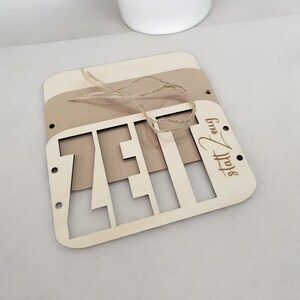 May include: A wooden sign with the word "Zeit" cut out in the center. The sign is made of light brown wood and has a brown paper backing. The sign is tied with twine and has the words "Stoff & Form" engraved in the bottom right corner.