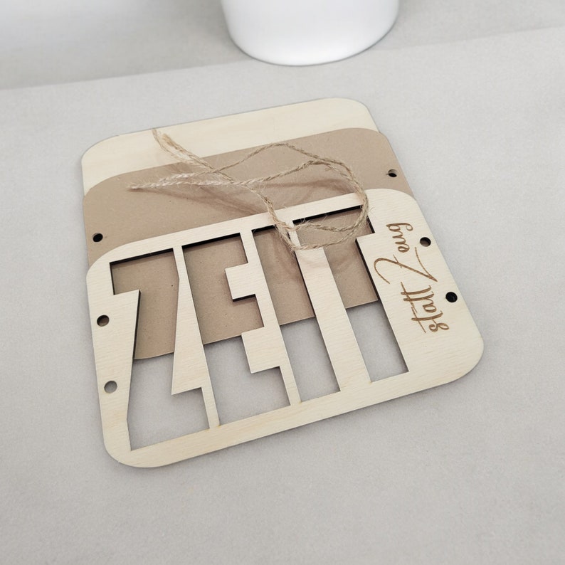 May include: A wooden sign with the word "Zeit" cut out in the center. The sign is made of light brown wood and has a brown paper backing. The sign is tied with twine and has the words "Stoff & Form" engraved in the bottom right corner.